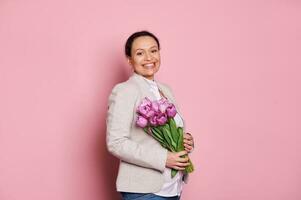 embarazada mujer sonriente un con dientes sonrisa mirando a cámara, participación un ramo de flores de tulipanes para De las mujeres día o de la madre día foto