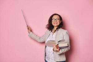 Positive pregnant female teacher in glasses, holding textbooks, smiling at camera, pointing with pointer at copy space photo