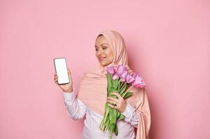 Muslim woman in hijab, with bouquet of tulips, looking at smartphone with white digital screen, ad space for mobile apps photo