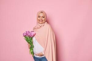 hermosa musulmán embarazada mujer con ramo de flores de tulipanes, conmovedor su barriguita, sonriente mirando a cámara, aislado en rosado foto