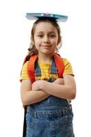 auténtico caucásico Chica de escuela con libro en su cabeza, sonriente a cámara, posando con brazos cruzado en blanco antecedentes foto