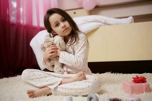 Adorable little girl hugs her plush toy, looking at camera, sitting on the carpet in her room. Children's Day. Childhood photo