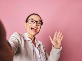 Selfie on smartphone of a stunning Latin American woman smiling and greeting with her hand, isolated on pink background photo