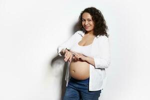 medio Envejecido embarazada mujer cheques móvil aplicación en inteligente mirar, cuenta bebé movimientos, sonriente a cámara, aislado en blanco foto