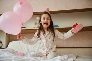 alegre cumpleaños niña en fiesta sombrero, participación manojo de rosado globos y regalo caja, sentado en cama y sonriente dentro cámara foto