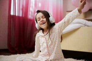 Adorable little girl, mischievous child dressed in pajamas, moving to music, singing, enjoying soundtrack on headphones photo