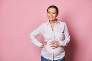 Attractive middle aged pregnant woman touching her belly, smiling looking at camera, enjoying a happy carefree pregnancy photo