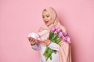 asombrado musulmán mujer en rosado hiyab, expresando felicidad, recepción un presente en corazón conformado regalo caja para de la madre día foto