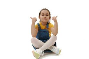 Lovely kid girl in casual denim clothes, looking at camera, gesturing with thumbs up, sitting on isolated white backdrop photo