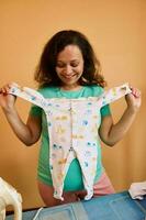 Smiling pregnant woman holding jumpsuit for newborn baby, standing by ironing desk, preparing bag for maternity hospital photo