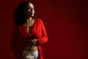 Attractive pregnant woman dressed in red wear, standing bare belly with a white orchid in hands, pensive looking away photo