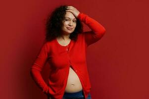 Confident portrait of beautiful pregnant woman, expectant mother, smiling looking at camera, isolated on red background photo