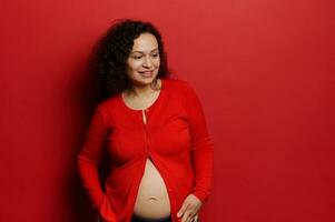 contento embarazada mujer en rojo desabrochado camisa, suavemente conmovedor su barriga, sonriente mirando aparte, aislado antecedentes. foto