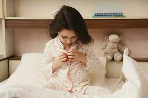 encantador pequeño niña botones arriba su pijama, se sienta en cama y obtiene Listo para cama. el concepto de yendo a cama y despertar foto