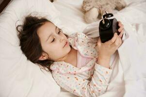 adorable pequeño niña en elegante pijama, acostado en cama debajo acogedor blanco funda Nordica, comenzando alarma reloj consiguiendo Listo para cama foto