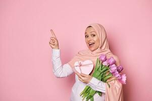 Delighted pregnant Muslim woman with a bouquet of tulips and a cute present, pointing at copy space on pink background photo