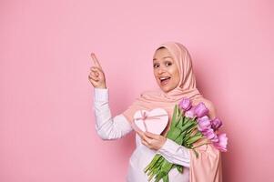 Delighted pregnant Muslim woman with a bouquet of tulips and a cute present, pointing at copy space on pink background photo