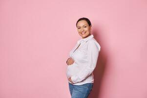 Multi ethnic pregnant woman, touching her belly, enjoying first baby kicks, smiles looking at camera. Healthy pregnancy photo