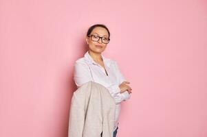 Serious businesswoman in glasses, looking confidently at camera, posing with her arms folded on isolated pink backdrop photo