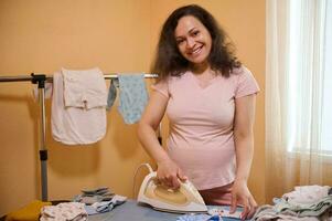 Middle aged multi ethnic positive smiling pregnant woman, future mom ironing baby clothes, preparing bag for child birth photo