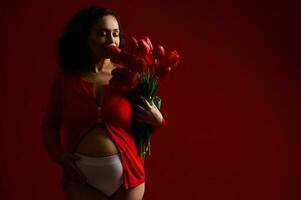 Gorgeous Latin American adult pregnant woman caressing her belly, sniffing a cute bouquet of red tulips for Mother's Day photo