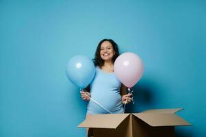 Beautiful pregnant woman holding blue and pink balloons, smiling looking at camera, isolated on blue color background photo