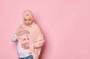 Medio este musulmán embarazada mujer, participación manos alrededor un rosado satín conciencia cinta en hiyab, aislado antecedentes foto