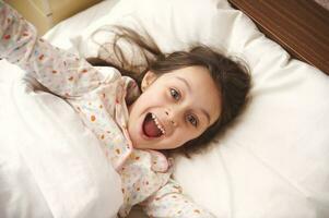 Mischievous little girl in pajamas, making faces at camera, stretching while waking up in morning, relaxing in her bed photo