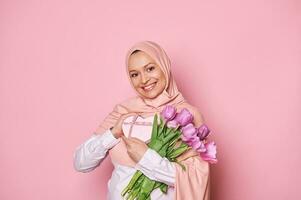 Positive Muslim woman with Mother's Day or birthday present and a bouquet of tulips, smiling on isolated pink background photo