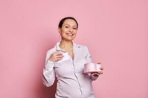 encantador maravilloso de edad mediana embarazada mujer con un regalo caja para de la madre día, sonrisas mirando a cámara, rosado fondo foto