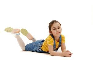 Beautiful Caucasian little schoolgirl in casual denim and yellow t-shirt, lying on white background, looking at camera photo