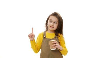 Lovely child girl holding cardboard mug with hot beverage, pointing finger at copy space on isolated white background photo