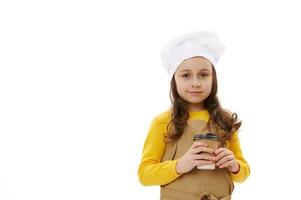 encantador niño niña en del chef sombrero y delantal, participación papel taza con para llevar caliente beber, aislado terminado blanco antecedentes foto