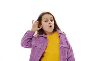 Curious little child girl puts hand on ear, eavesdrops a conversation, looks at camera with open mouth, white background photo