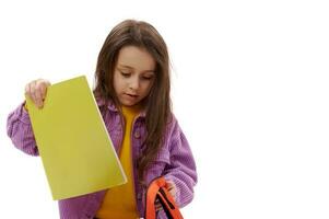 Close-up adorable elementary age student, cute kid girl putting textbook into her backpack, isolated on white background photo