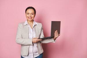 Mature business woman smiling, confidently looking at camera, holding laptop isolated on pink background. People concept photo