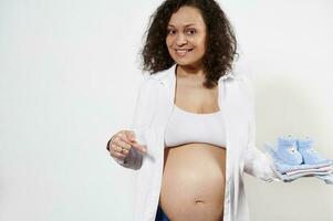 Charming curly haired middle aged gravid woman holding newborn clothes, smiling pointing at her belly, isolated on white photo