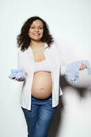 Cheerful pregnant woman holding blue baby booties and bodysuit for her future newborn child, smiling on white background photo