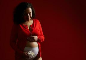 delicia maravilloso adulto embarazada mujer sonriente, acariciando su barriga, posando con un blanco orquídea flor, rojo antecedentes foto