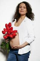 latín americano embarazada mujer participación rojo tulipanes, acariciando su barriga, sonriente mirando a cámara. el embarazo 28 semana foto