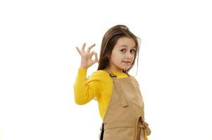 Caucasian kid girl in chef's apron, little waitress in child cafe, gestures with OK sign, expressing positive emotions photo