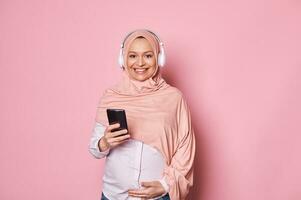 Smiling pregnant Muslim woman in pink hijab and headphones, checking mobile application on smartphone, smiling at camera photo