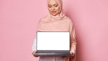 Focus on laptop with white blank screen in the hands of Arab Muslim pretty woman in hijab, isolated on pink background photo