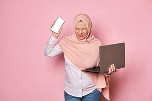 Pregnant Muslim woman in hijab, with smartphone and laptop, overworked with office job, looks forward to maternity leave photo