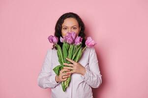 Portrait of a beautiful woman picking out from a bouquet of purple tulips, looking at camera on isolated pink background photo