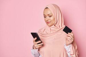 hermosa Arábica musulmán mujer en rosado hiyab, utilizando teléfono inteligente, participación crédito tarjeta, haciendo pago vía Internet bancario foto