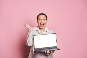Surprised multi ethnic woman with her mouth open, holding laptop with white blank screen, isolated on pink background photo