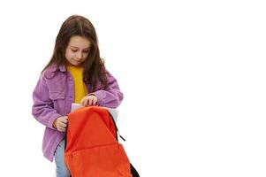 Adorable schoolgirl folds her school supplies in her backpack, sitting over isolated white background. Childhood. School photo