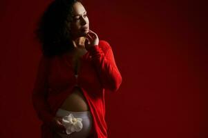 Beauty portrait of ethnic pregnant adult woman with white orchid flower, posing bare belly, isolated on red background photo