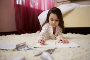 Lovely little girl in pajamas, lying down on carpet in bedroom, concentrated on drawing picture with colorful pencils photo